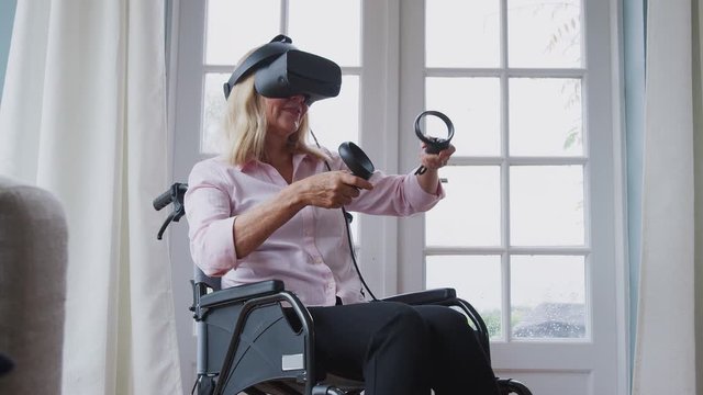 Mature Disabled Woman In Wheelchair At Home Using Virtual Reality Headset Gaming Holding Controllers