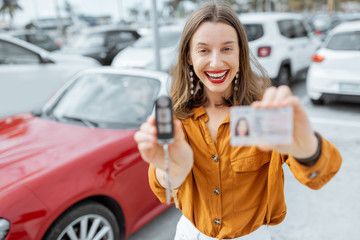 Portrait of a happy woman showing driver's license and keys standing near the red cabriolet at the...