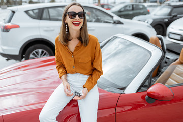 Portrait of a cheerful young woman near the red cabriolet at the car parking outdoors. Happy car ownership concept