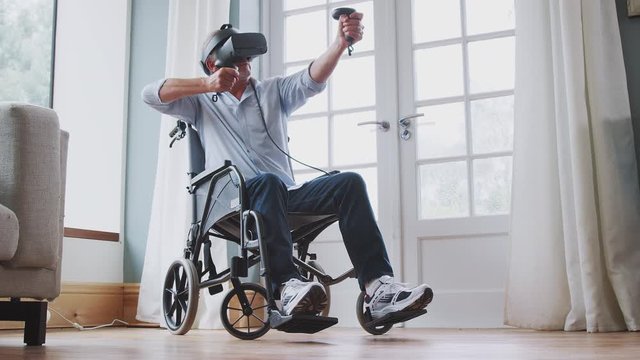 Senior Disabled Man In Wheelchair At Home Wearing Virtual Reality Headset Holding Gaming Controllers