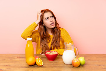 Fototapeta premium Teenager redhead girl having breakfast in a table having doubts and with confuse face expression