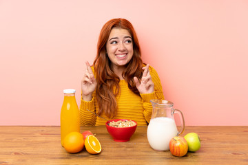 Teenager redhead girl having breakfast in a table with fingers crossing