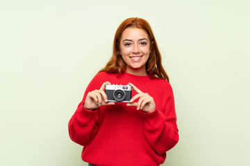 Teenager redhead girl with sweater over isolated green background holding a camera