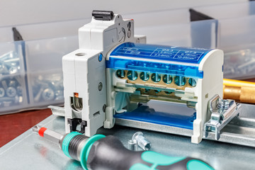 Installed on DIN rail on metal mounting plate automatic circuit breaker and wire terminal against transparent plastic box with screws and nuts and screwdriver on foreground