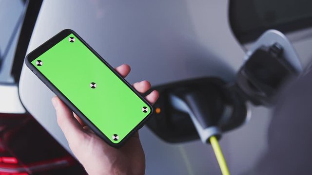 Man Charging Electric Vehicle With Cable Looking At App On Mobile Phone