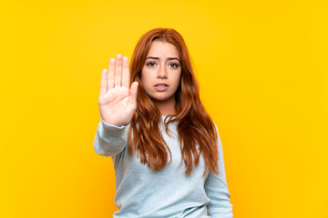 Teenager redhead girl over isolated yellow background making stop gesture