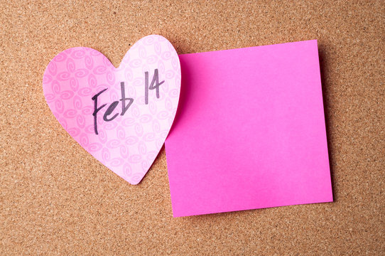 Handwritten reminder of Feb 14th Valentine's Day on pink heart with sticky note copy space on bulletin board 
