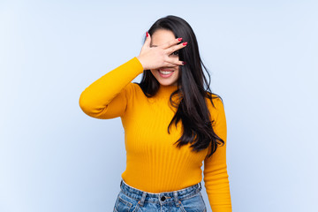 Young Colombian girl over isolated blue background covering eyes by hands and smiling