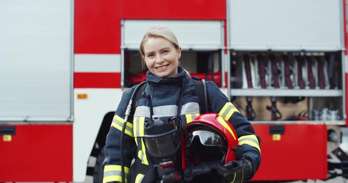 Portrait shot of the young blonde caucasian woman fire fighter in her full equipment standing at the big car and smiling.