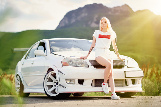 Beautiful, sexy young blonde with tattoos in short shorts and a T-shirt sits on the hood of a white sports car on a background of mountains