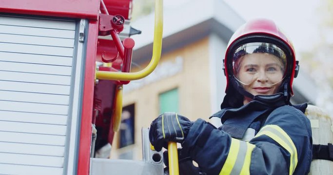 Close up of the blonde young Caucasian female fire fighter going up the ladder on the fire truck and then smiling to the camera. Portrait.