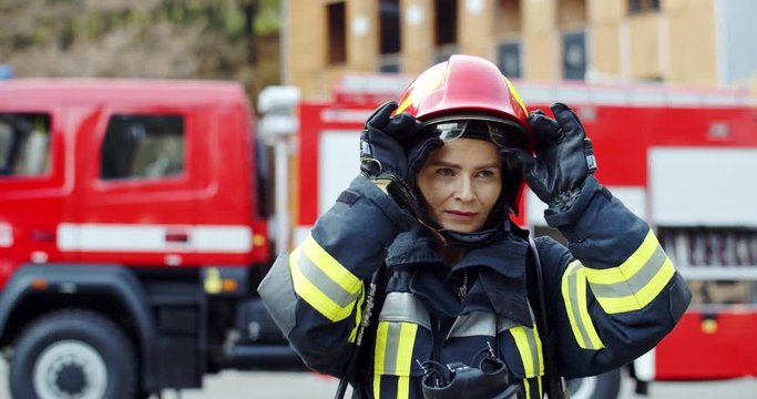 Portrait of the beautiful Caucasian female fireguard in the equipped costume and helmet taking off goggles and smiling to the camera. Outdoor.