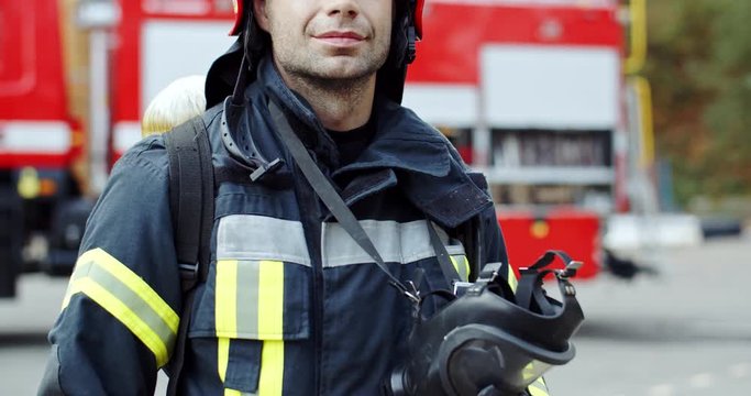 Portrait shot of the good looking Caucasian young fireman in the helmet and with a gas mask in hands looking and smiling to the camera. Outside.