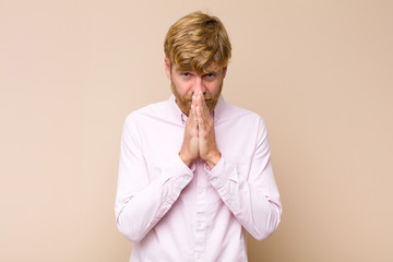 young blonde man feeling worried, hopeful and religious, praying faithfully with palms pressed,...