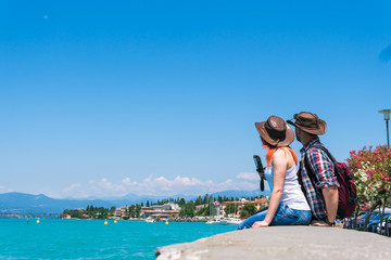 Fototapeta na wymiar A happy couple in a cowboy hat and a backpack sits on the promenade, looks at the turquoise water of Lake Garda and use smartphone. Vacation and tours to Europe. Sunny summer day. Italy