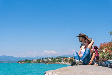 Fototapeta na wymiar A couple in a cowboy hat and a backpack sits on the promenade, looks at the turquoise water of Lake Garda. Vacation and tours to Europe. Sunny summer day. Italy. Mountains background, copy space