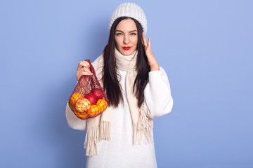 Horizontal shot of young woman holding shopping bag full of apples and oranges, dresses white warm...