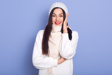 Picture of adorable magnetic energetic young lady with black long hair and red lips, raising one hand, putting hand on cheeck, smiling sincerely, being in high spirits, wearing warm clothes.