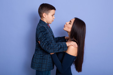 Indoor picture of lovely adorable mother and little son, going to kiss each other, spending time together, holding each other, wearing dark blue items of clothes. Family relationship concept.