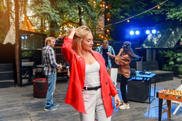 Close up of attractive blond girl with blue eyes posing to the camera with her friends dancing in disco light on the background. Outdoors evening party concept.