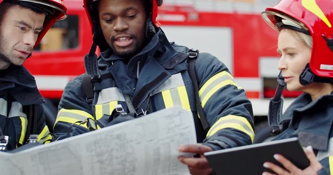 Two multiethnic men firefighters in helmets with a building plan or map in hands deciding how to fight with a fire and female colleague with tablet device listening to them.