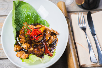 Sliced chicken fried in sweet and sour soy sauce with vegetables and herbs in a deep plate on a wooden table in a restaurant.