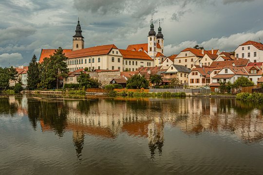 Telc / Czech Republic - September 27 2019: View of the castle, the tower of the church of James the Great and historical houses over a lake. Reflection of the cityscape and dramatic blue sky in water.