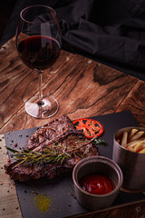 Cooked meat t-bone steak on serving board with garlic cloves, tomatoes, rosemary, spices and glass of red wine over rustic wooden background