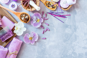 Top view of violet purple set for relaxing spa treatments. Wooden mortar pounder with herbs, spoons with spices, candles, aroma sticks, towel rolls, handmade soap, bath bomb are on cement background.