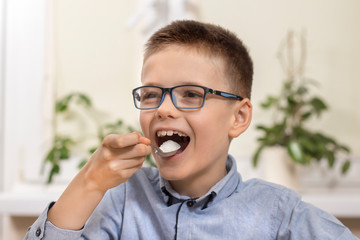 Uśmiechnięty chłopiec w wieku szkolnym siedzi przy stole i wkłada do ust łyżeczkę z białym cukrem. - obrazy, fototapety, plakaty