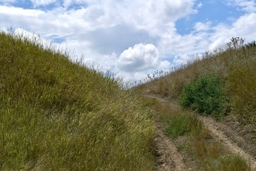 The landscape is a rustic view of a country road in the center between the hills and the blue cloudy sky.