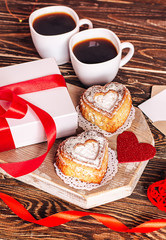 Romantic breakfast for Valentine's Day. Two cups of coffee, cupcakes in the form of a heart on a white board on a wooden table, gift with a red ribbon. Copy space, close up