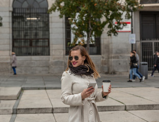 Attractive slim blonde girl with her mobile phone and a takeaway coffee smiles on a city street