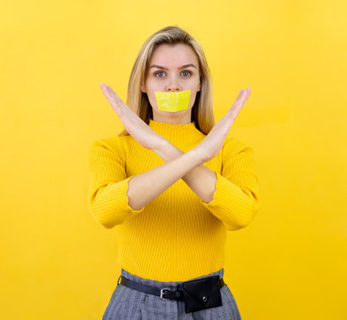 Portrait of a blonde girl with crossed hands gesture and yellow tape taped her mouth. Censorship concept on yellow background. Stop, warning