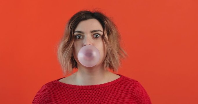 Portrait happy girl inflates bubble from chewing gum balloon and bursts it in red sweater on red background smiling looking at camera. Monotone. Slow motion. Emotions of people. Lifestyle