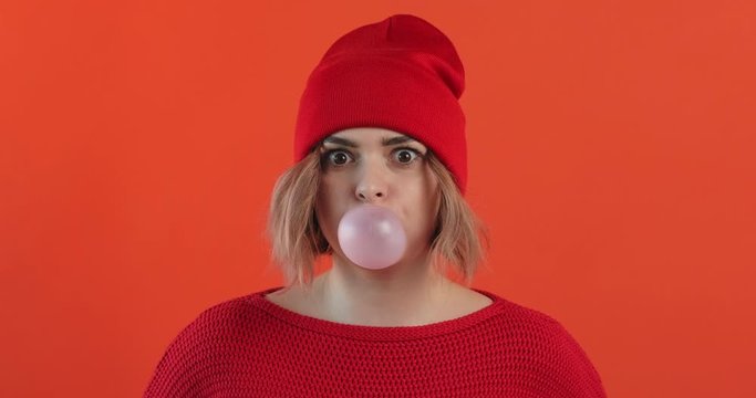 Portrait happy girl in red hat inflates bubble from chewing gum balloon and bursts it in red sweater on red background smiling looking at camera. Monotone. Slow motion. Emotions of people. Lifestyle