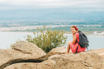 Young girl resting on the top of mountain, admiring sea landscape, wearing backpack, listening music with headphones
