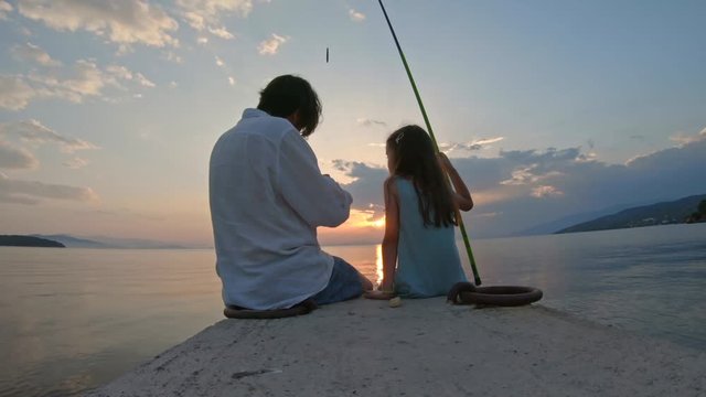 Rear view of little girl sitting on the edge of a dock and fishing with father Mediterranean Sea at sunset. Enjoying adventure in summer vacation	