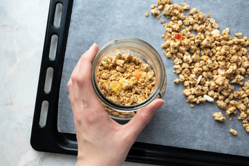 Hand holding glass jar with granola on parchment paper on baking sheet, top view. Made homemade healthy breakfast