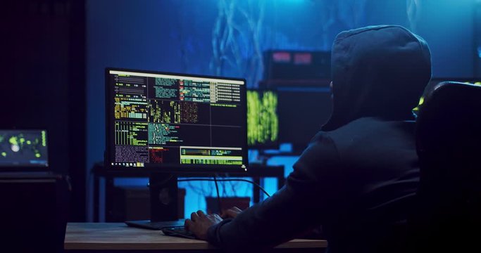 Back view on the man in a hood sitting and working at the computer in the dark room with technologies, hacking the system and laundring data. Rear.