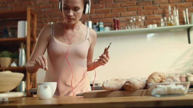 Young woman dunking sugar in coffee. Dancing girl listening music in kitchen.