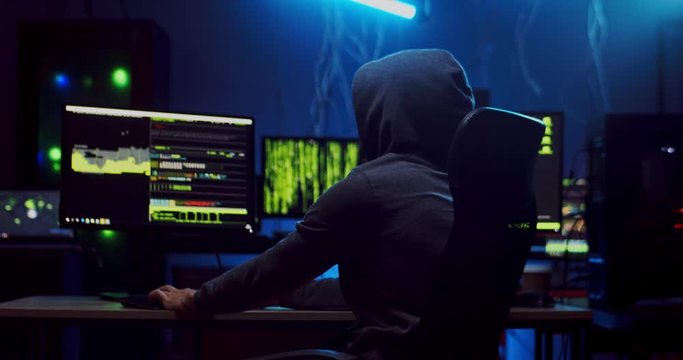 Rear of the anonymous male hacker in a hood coding a virus or secret program in the software dark room. Back view.