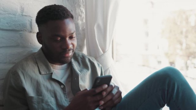 Close up view of handsome smiling african american young man relaxed sitting on windowsill in apartment with beautiful city views holding phone and surfing in the Internet