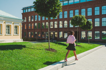 back view of unrecognizable young girl in a green skirt, pink jacket and sports sneakers who walks in the city holding a paper Cup of coffee in her hand