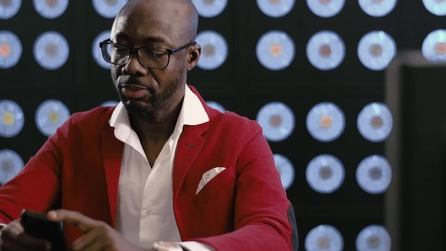 Cute mature bald african-american businessman, in red suit, white shirt and stylish glasses uses smartphone holding it in hands, sitting in dark room with blurred background having blue classic color.