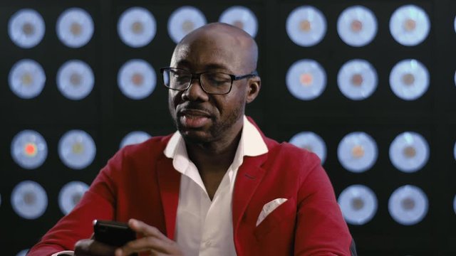 Smiling bearded bald african-american businessman, in red suit, white shirt and stylish glasses looks at smartphone holding it in hands, sitting in dark office with background of blue classic color.