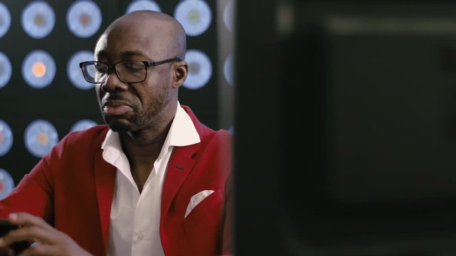 Attractive bearded bald african-american businessman, in red suit, white shirt and stylish glasses looks at smartphone holding it in hands, sitting in dark office with background of blue classic color
