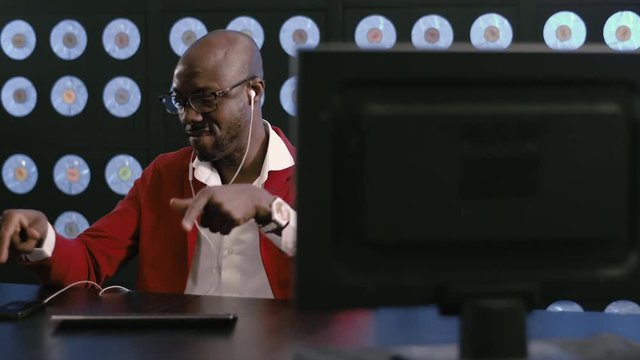 African-american bearded bald guy, wearing red suit, white shirt, stylish glasses and white phone headset, works on computer listening to music, sitting in dark room bluse classic background.