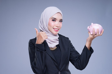 Finance and accounting savings concept.  A Muslim woman in business attire with a piggy bank isolated on a grey background.