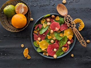 Top view of mixed citrus fruit salad, with spinach leaves and hazelnuts in a black dish with a wooden spoon. Mixed tropical fruits (lime, lemon, orange, grapefruit, mandarin), summer, colorful salad